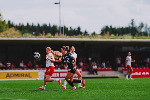Fußballerinnen im Zweikampf um den Ball auf einem Rasenplatz mit Zuschauern im Hintergrund