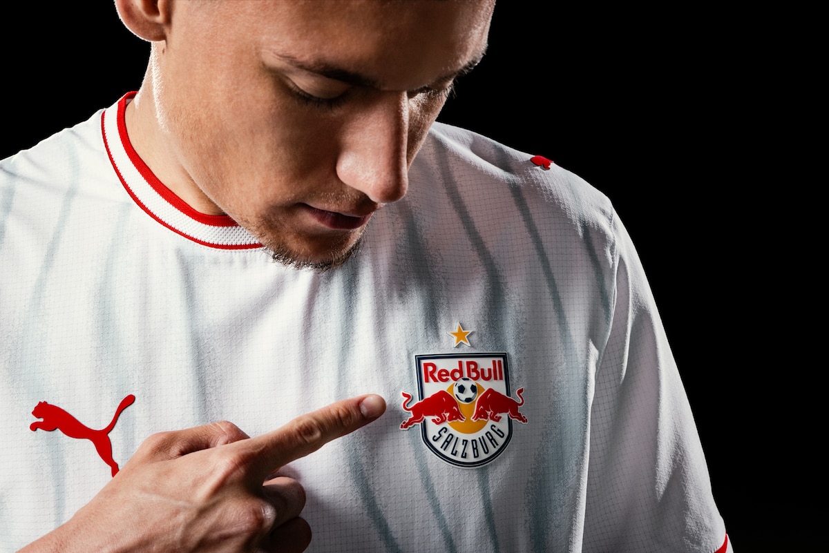 Fußballer zeigt auf das Wappen des FC Red Bull Salzburg auf einem weißen Trikot.