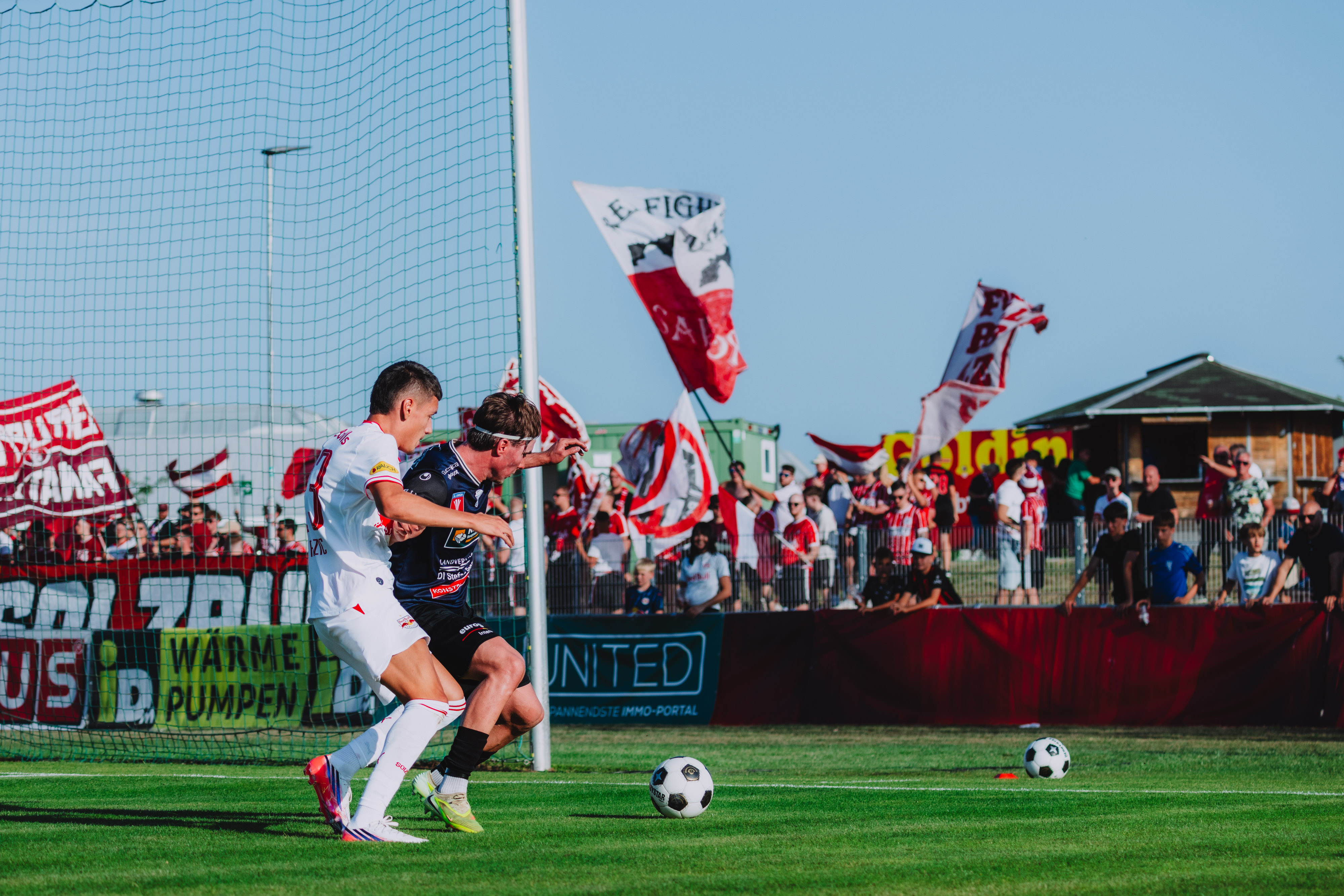 Fußballer kämpfen um den Ball, Fans mit Fahnen im Hintergrund auf einem Rasenplatz.