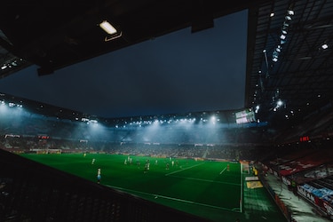 Fußballstadion bei Nacht mit Spielern am grünen Rasen und Flutlichtstrahlen.