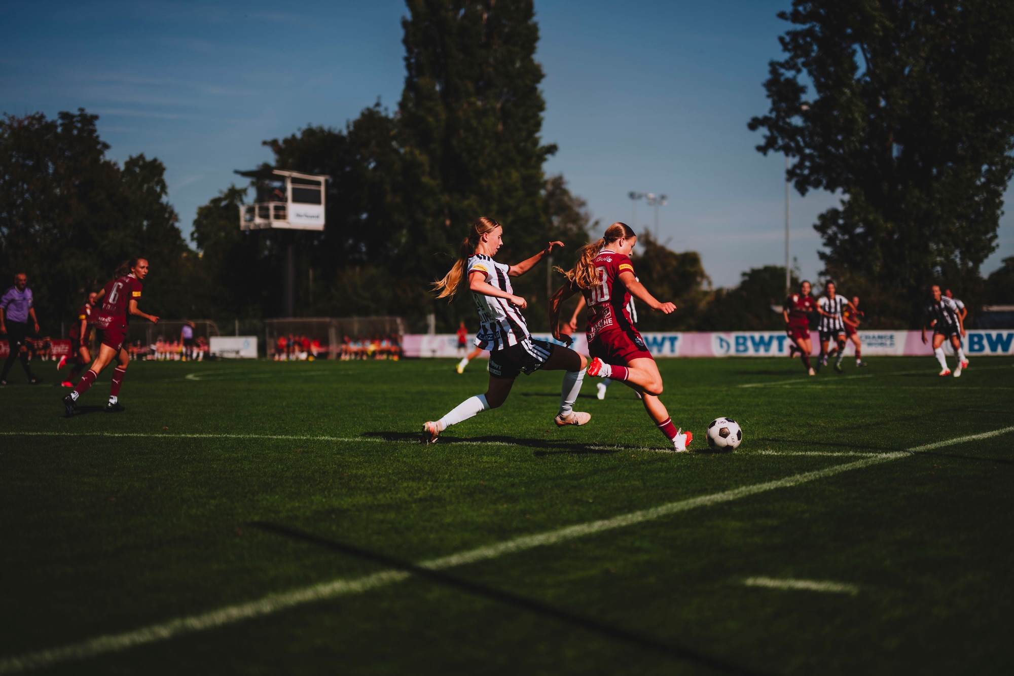 Fußballspielerinnen im Spiel auf grünem Rasen, mit Bäumen und Tribüne im Hintergrund